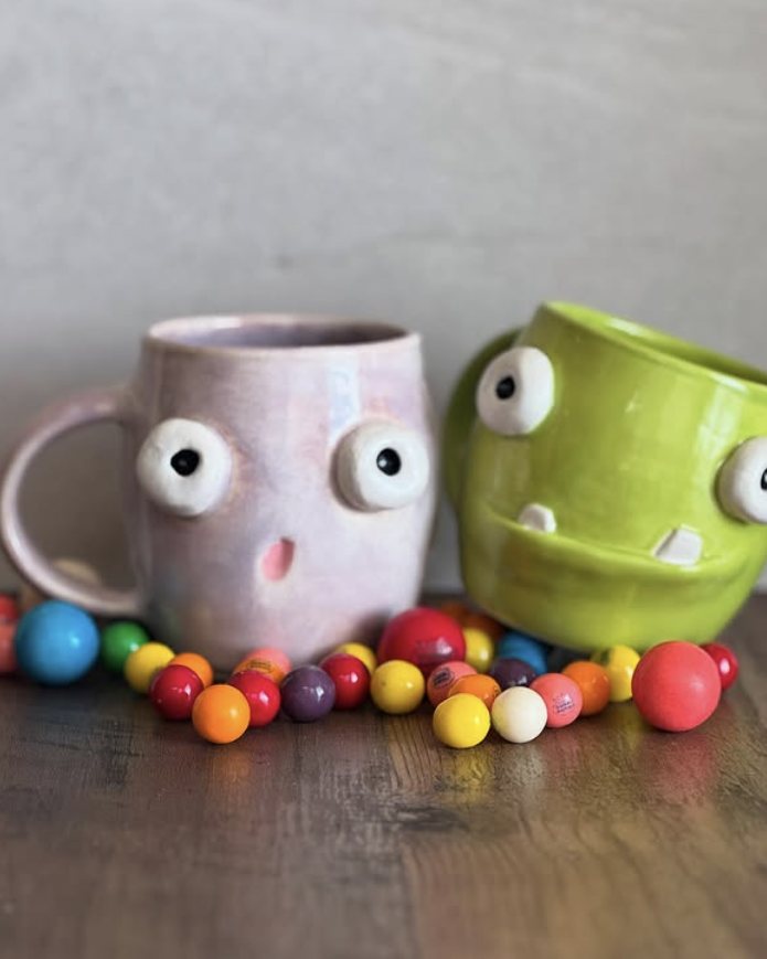 Two handmade mugs with googly eyes, one pale pink and one lime green, surrounded by gumballs